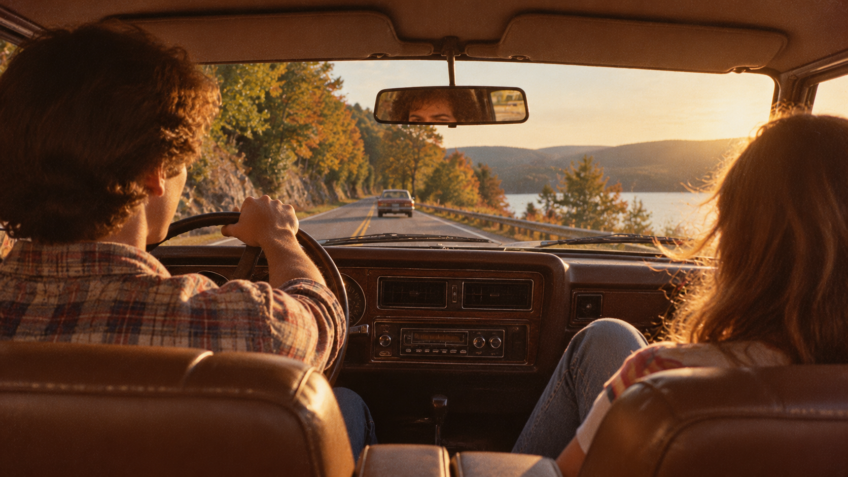 Two people drive along a road beside a lake during sunset, viewed from the backseat of the car.