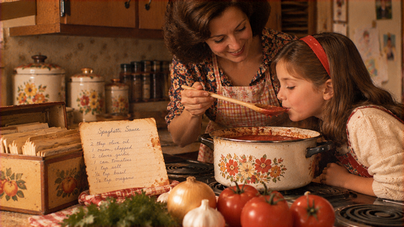 A woman and young girl taste spaghetti sauce from a wooden spoon held over a floral-patterned pot, with a handwritten recipe card displayed on the counter.