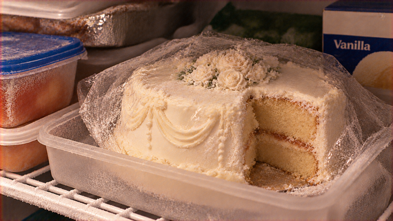 A frosted white layer cake with a slice removed, stored in a plastic container inside a freezer.