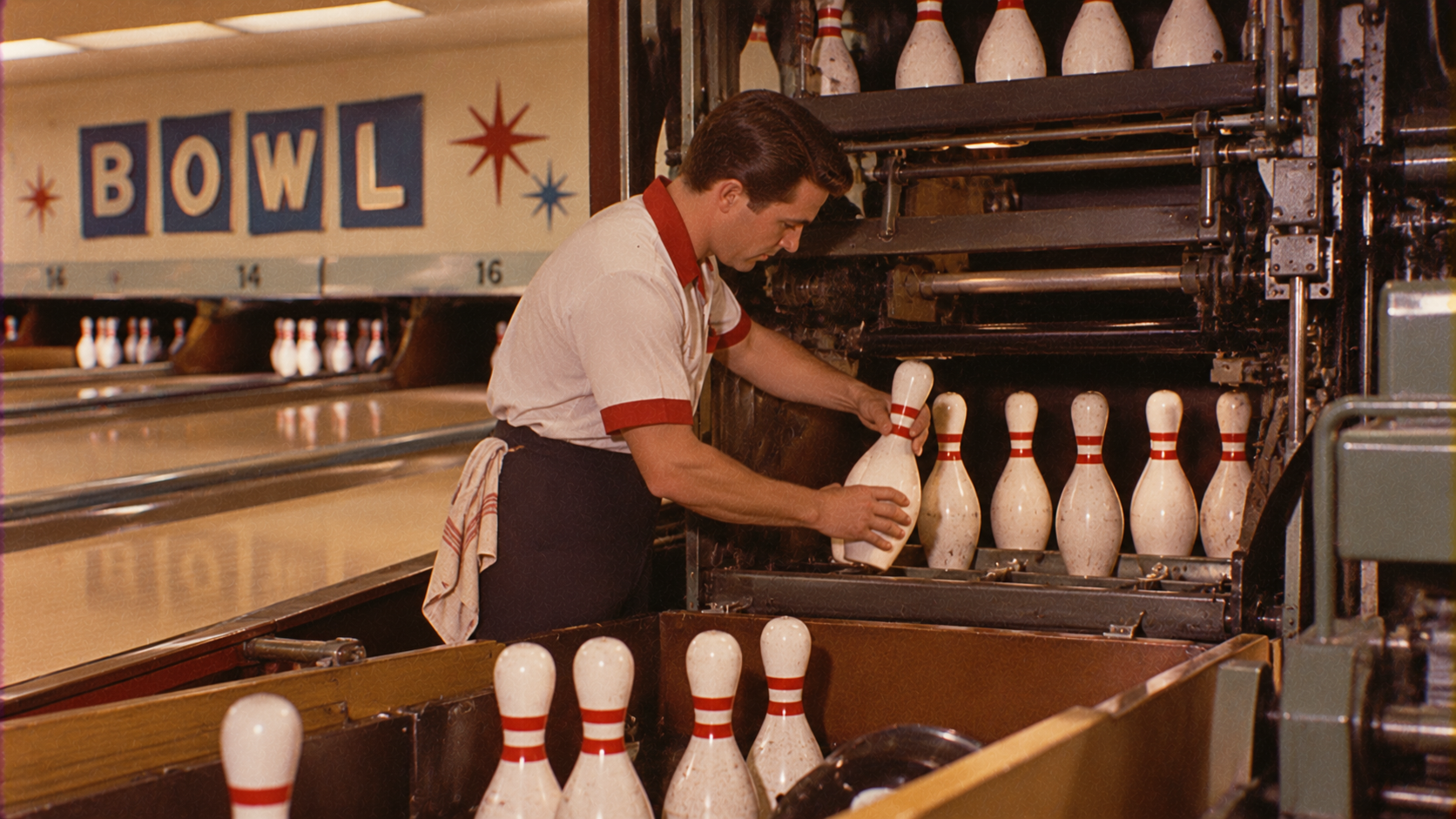 A man in a bowling alley, wearing a white and red shirt, resets bowling pins by hand in a pinsetter machine. The word “BOWL” is visible on the wall above the lanes in the background.