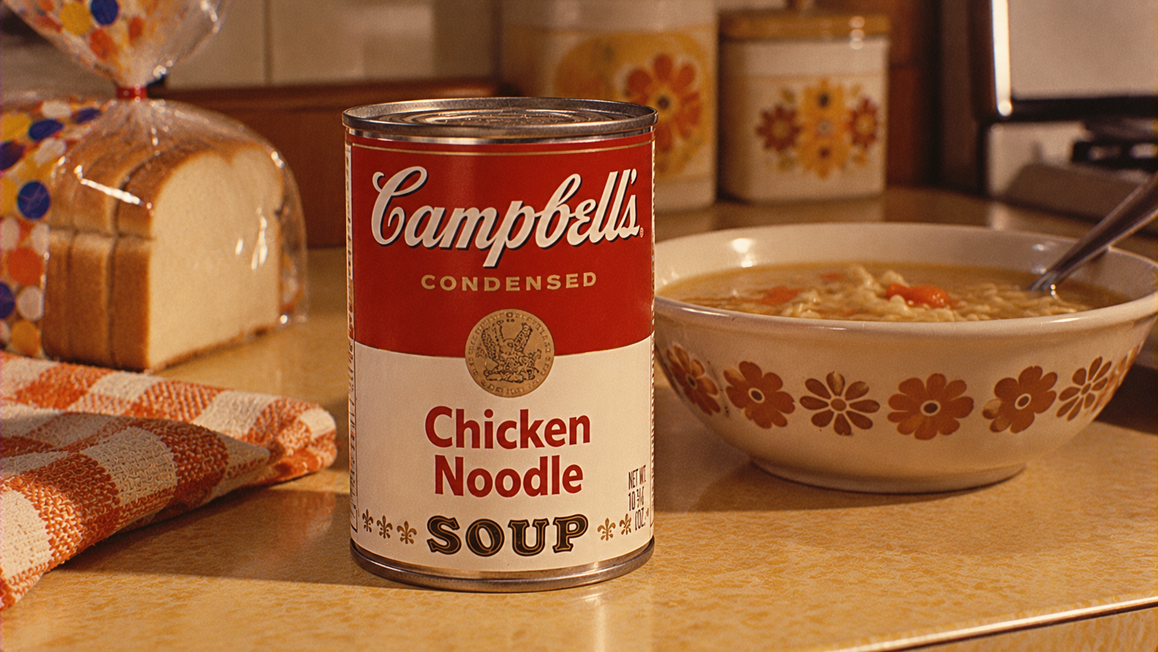 A can of Campbell’s Chicken Noodle Soup sits on a kitchen counter next to a bowl of soup, a loaf of bread, and a checkered towel.