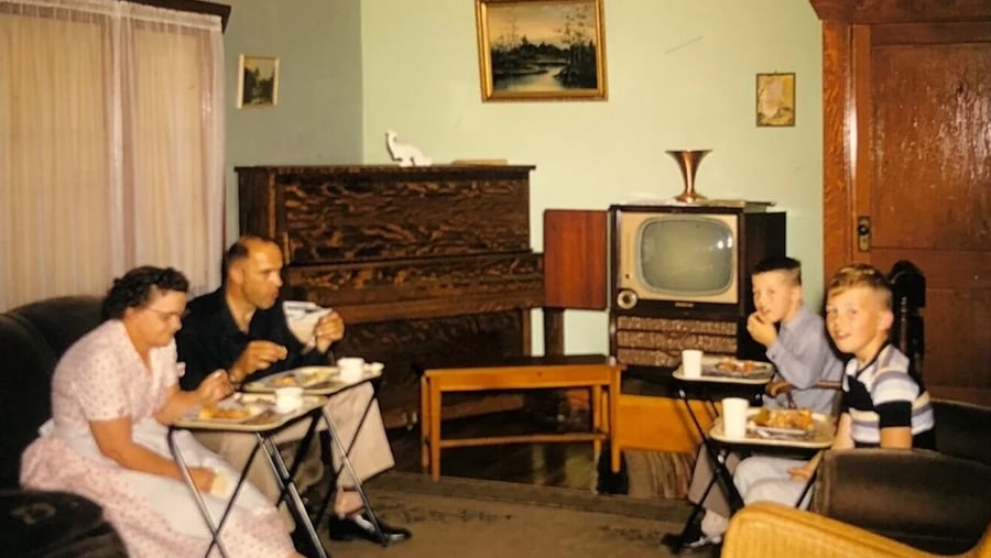 A family of four eats dinner on TV trays in a vintage living room with an old television, piano, and paintings on the wall.