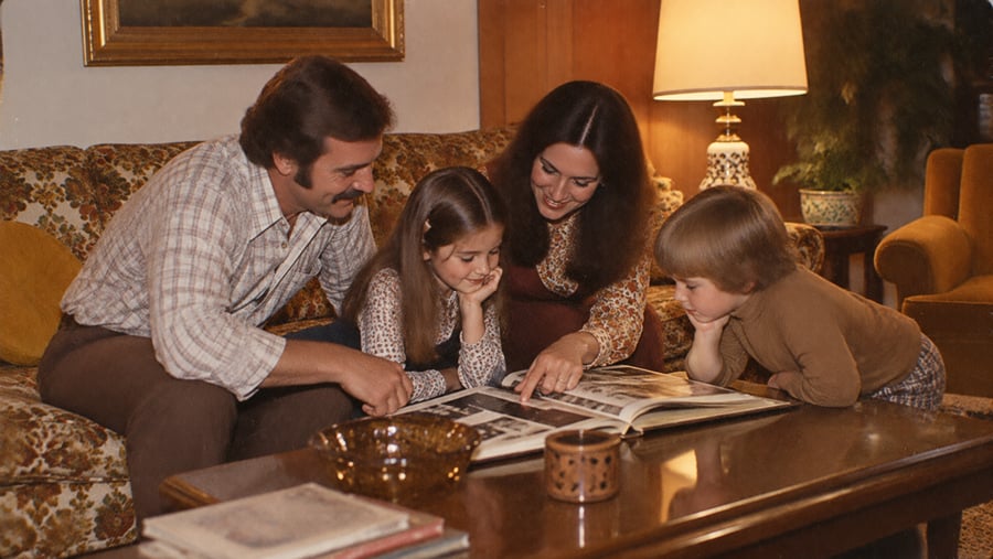 A family of four sits on a couch, smiling and looking at a photo album together on a coffee table in a cozy, warmly lit living room.