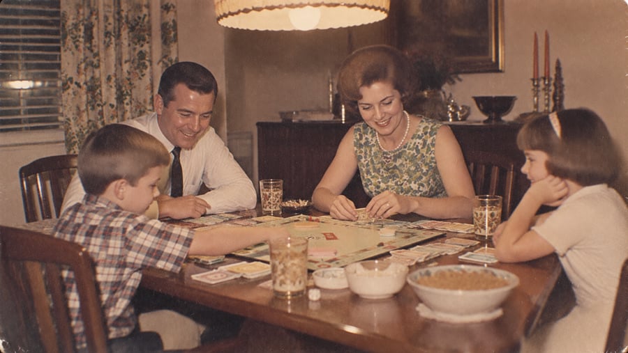A family of four sits around a dining table playing a board game, smiling and enjoying themselves. Drinks and snacks are on the table; a lamp hangs overhead, giving the scene a warm, cozy atmosphere.