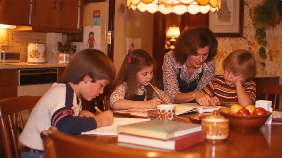 A woman helps two children with homework at a wooden table, while another child writes. The table has books, mugs, and a fruit bowl. The setting is a cozy kitchen with warm lighting.