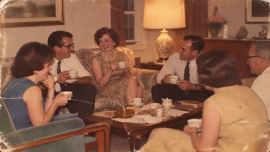 Six adults in vintage clothing sit around a coffee table, smiling and drinking tea in a cozy living room with warm lighting.