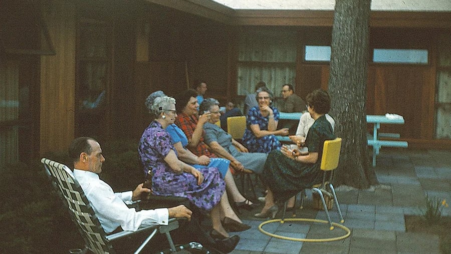 A group of older adults sit and talk in a shaded patio area. Some are in dresses, and one man in a white shirt relaxes in a lawn chair. Yellow chairs and a hula hoop are visible. The setting appears calm and social.