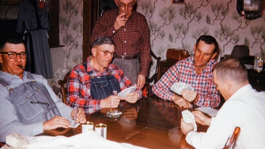 Five men, some in plaid shirts and overalls, sit around a wooden table playing cards. Two smoke pipes, and tin cans and snacks are on the table. The scene appears casual and vintage, with patterned wallpaper in the background.