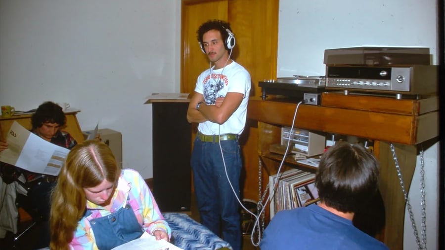 Four people are in a room with music equipment and papers. One person stands wearing headphones, while three others sit reading. The scene appears casual and focused, with records and a record player visible.