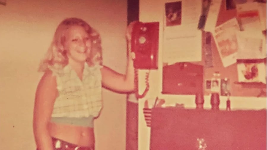 A woman smiles while holding the receiver of a red rotary wall phone. She wears a sleeveless plaid crop top and stands next to a bulletin board with various papers. The photo has a vintage, faded look.
