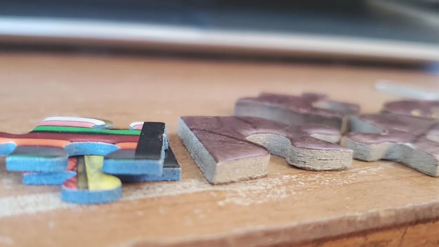 Close-up of two jigsaw puzzle pieces on a wooden surface, one with a colorful pattern and the other with a muted brown design, both lying flat with visible texture.