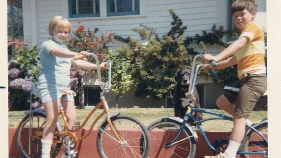 Two children on bicycles smile at the camera, with a black dog sitting between them. They are outside, in front of a house with plants and a red brick wall.