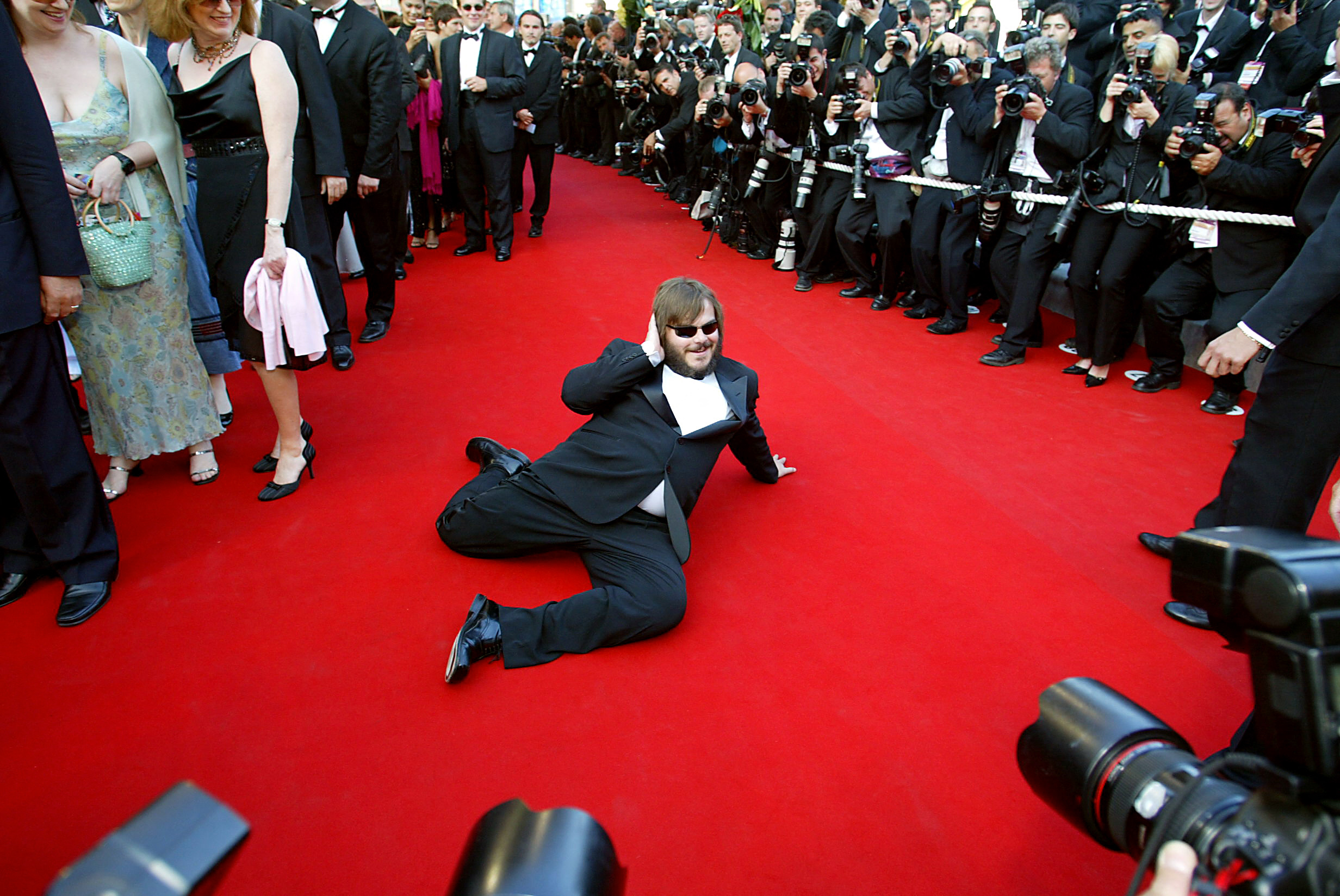 Jack Black sits on a red carpet, posing for a large group of photographers.