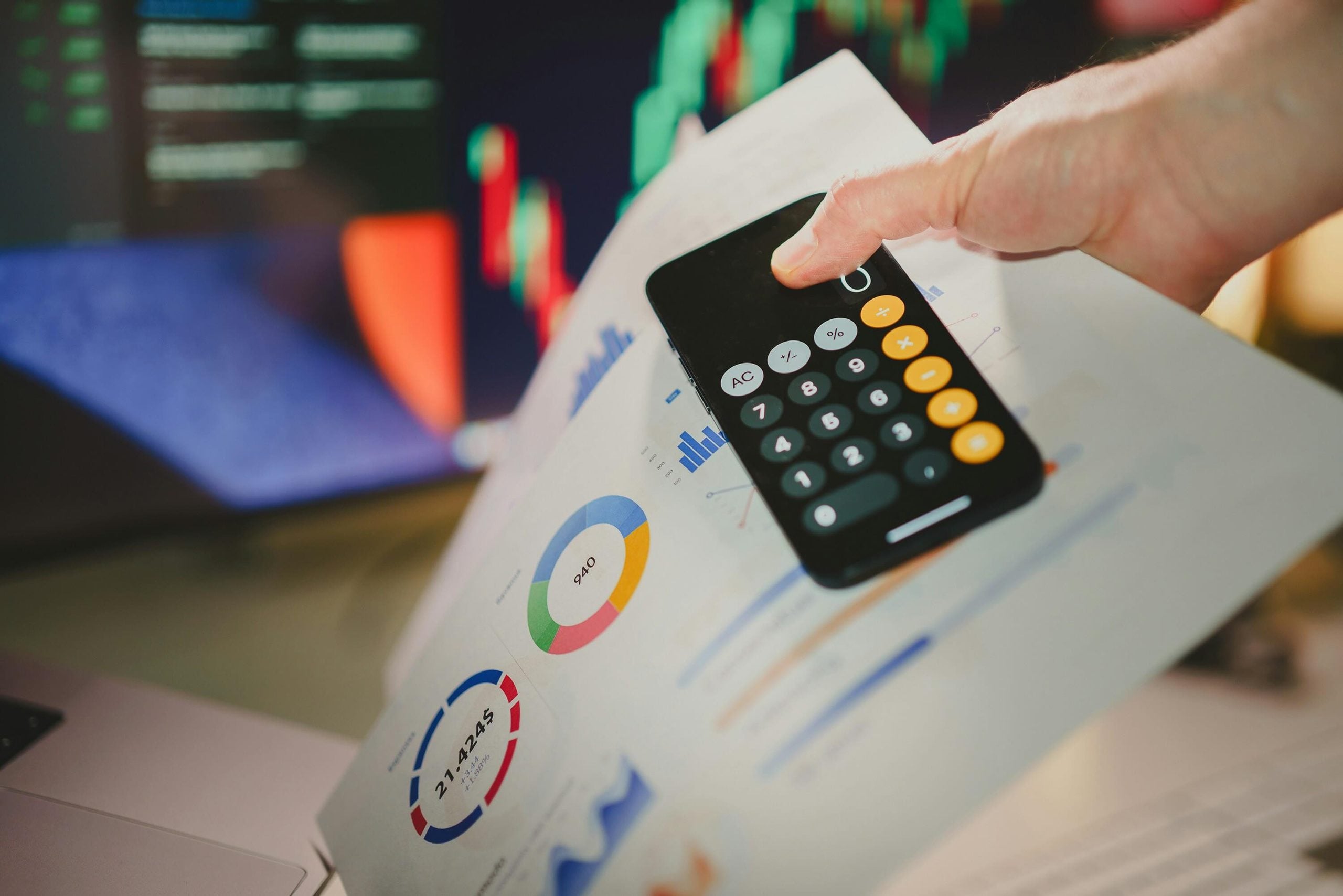 A hand holds a smartphone displaying a calculator app over a printed document with financial charts and graphs.