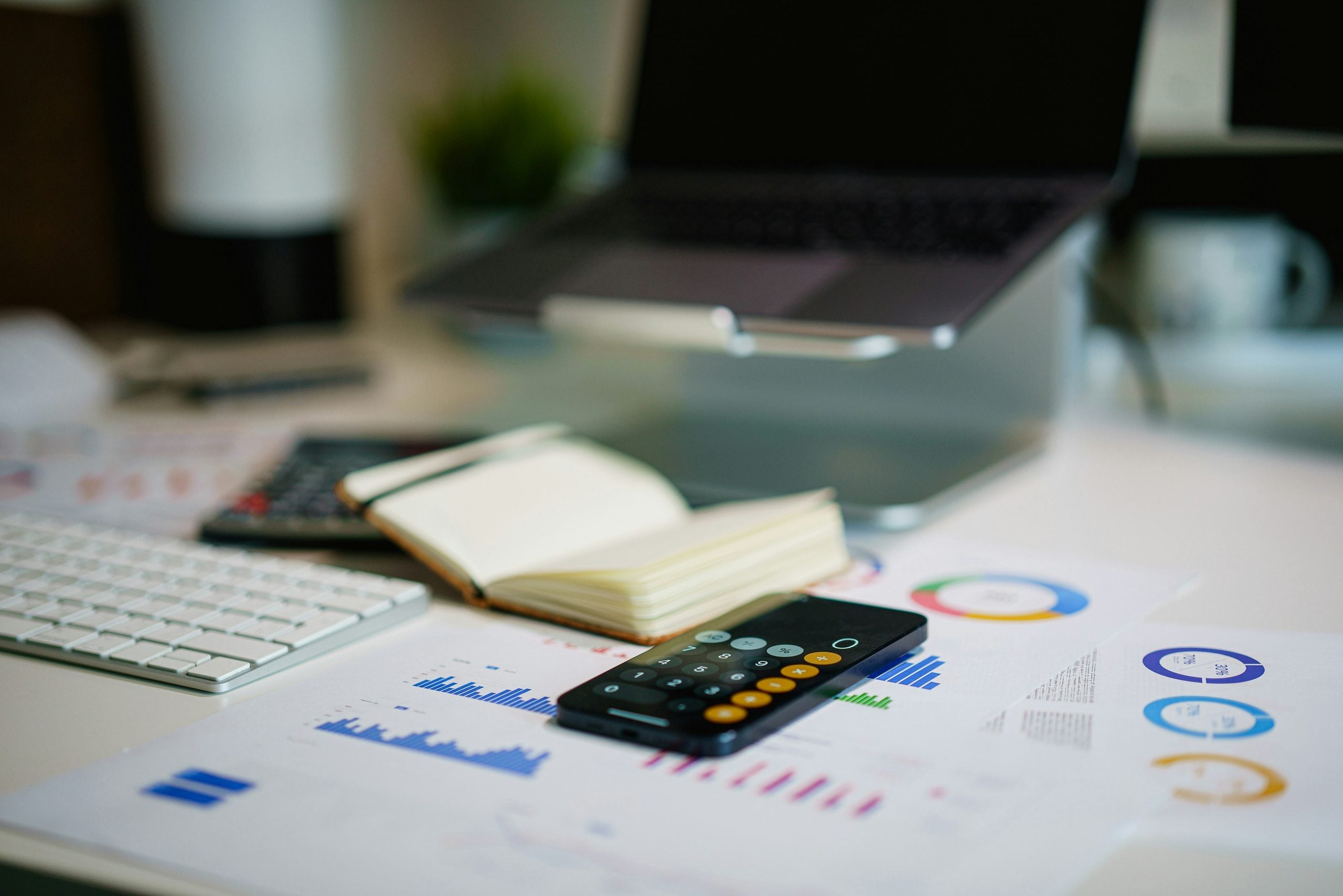 A smartphone displaying a calculator app rests on a desk cluttered with financial charts, an open notebook, and a laptop.