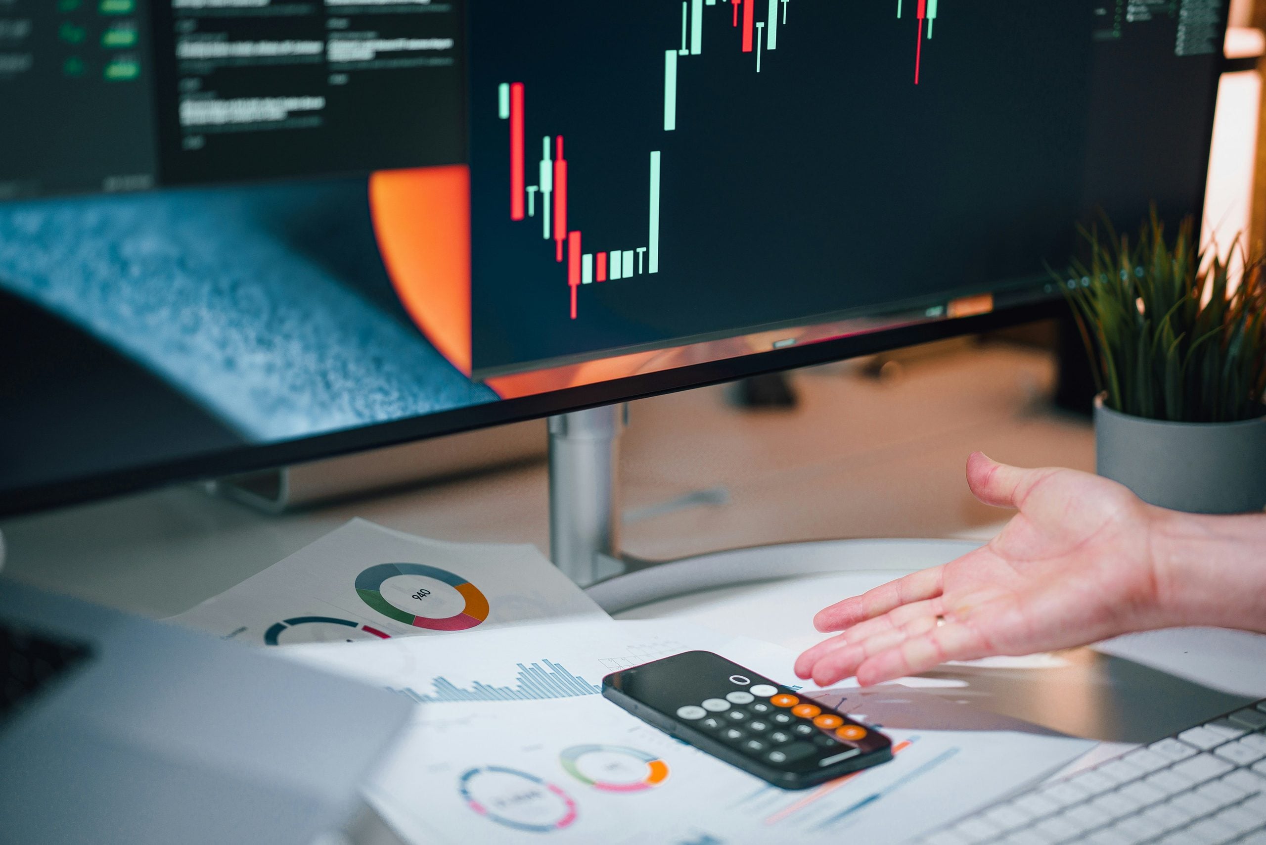 A hand gestures toward a desk featuring financial charts on a computer monitor, a calculator, and papers with printed data graphics.