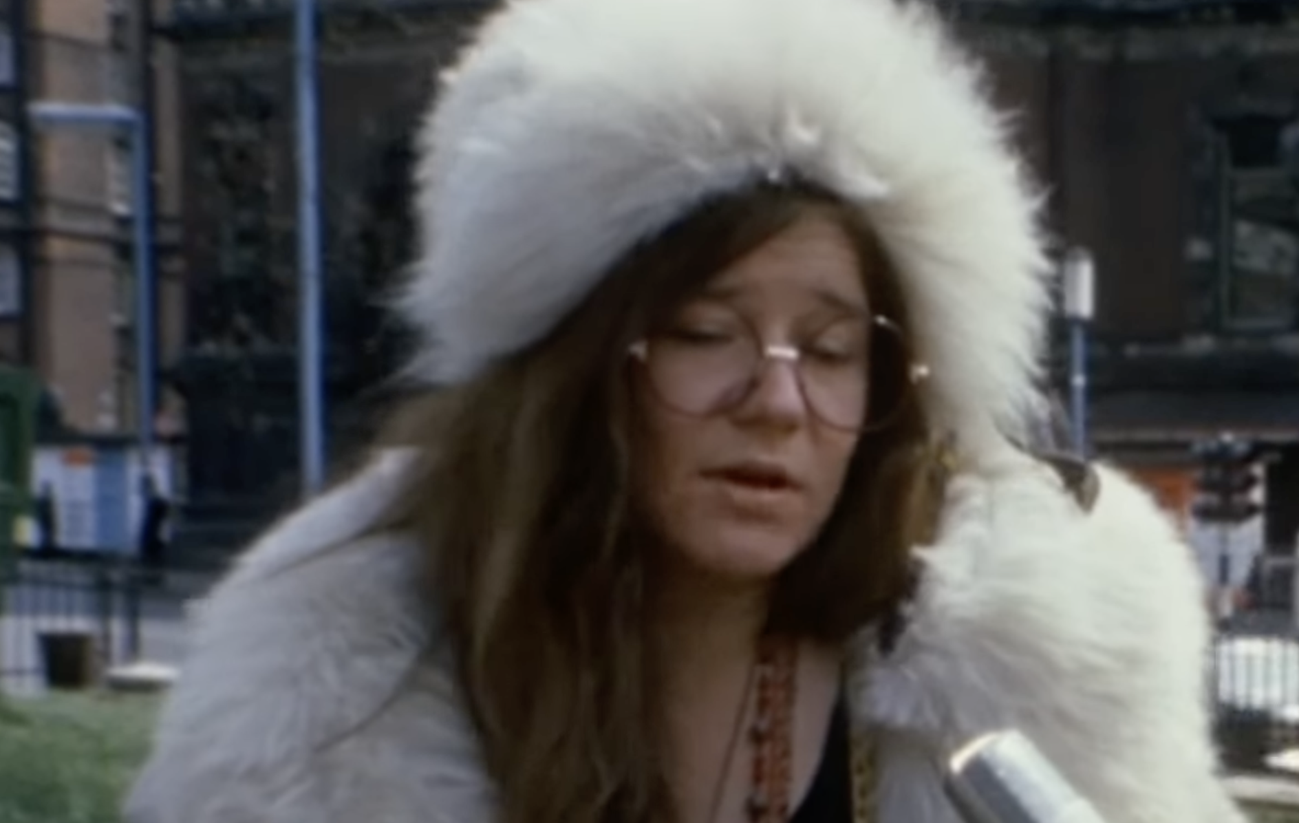 Janis Joplin wearing a large white faux fur hat and coat while speaking into a microphone.