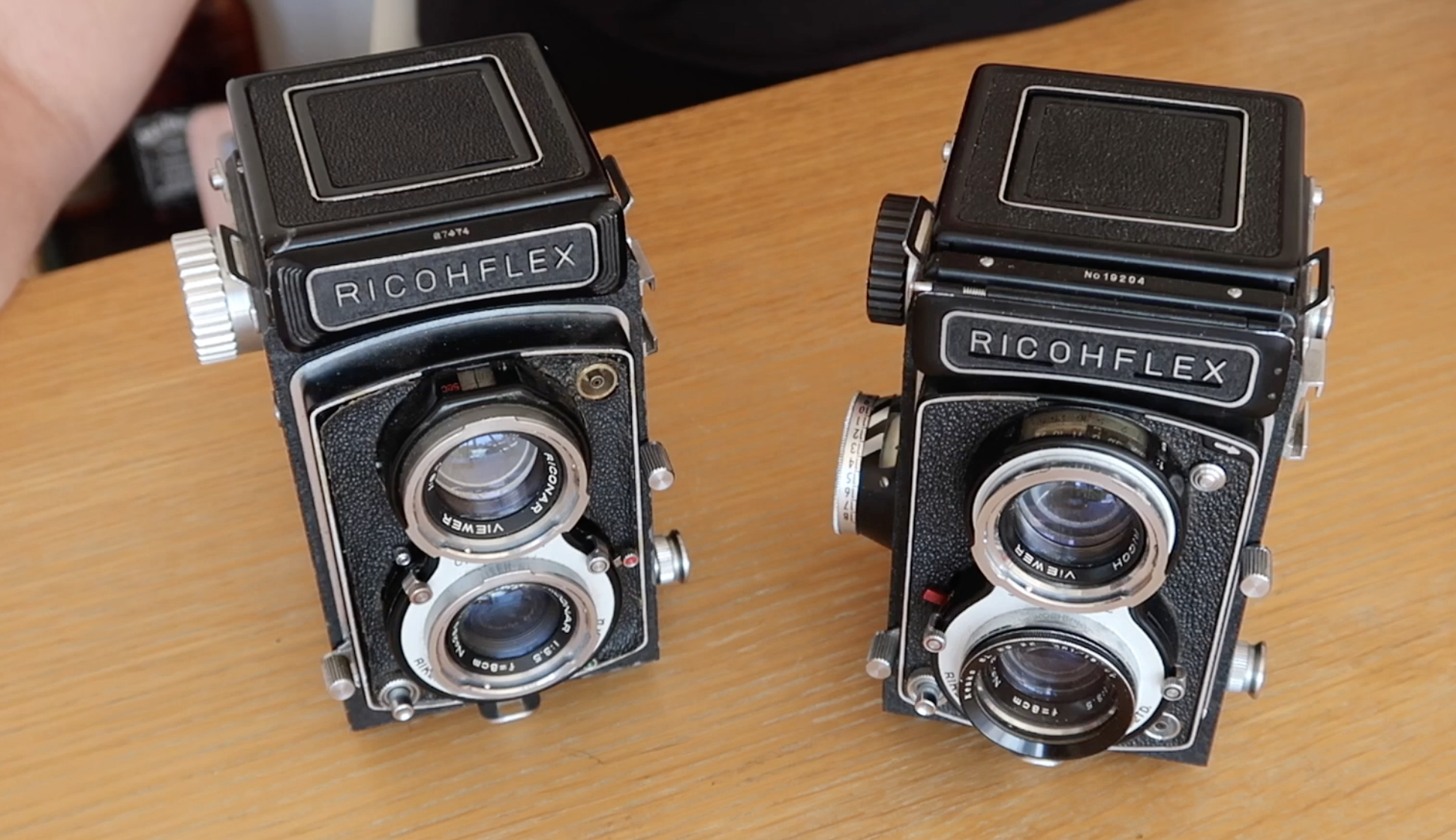 Two vintage Ricohflex twin-lens reflex cameras sitting on a wooden surface.