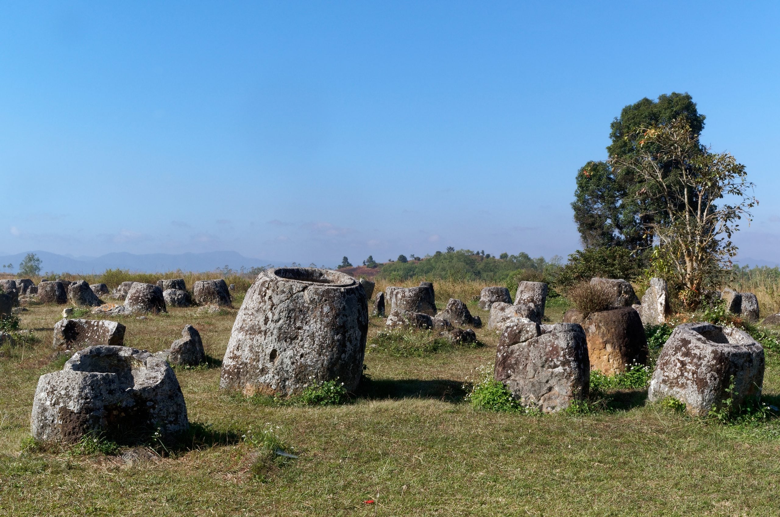 Ancient stone megalithic jars scattered across a grassy hillside under a clear blue sky.