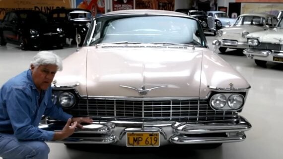 A man in a blue shirt kneels next to a classic pink Cadillac with a yellow license plate in a showroom filled with other vintage cars.