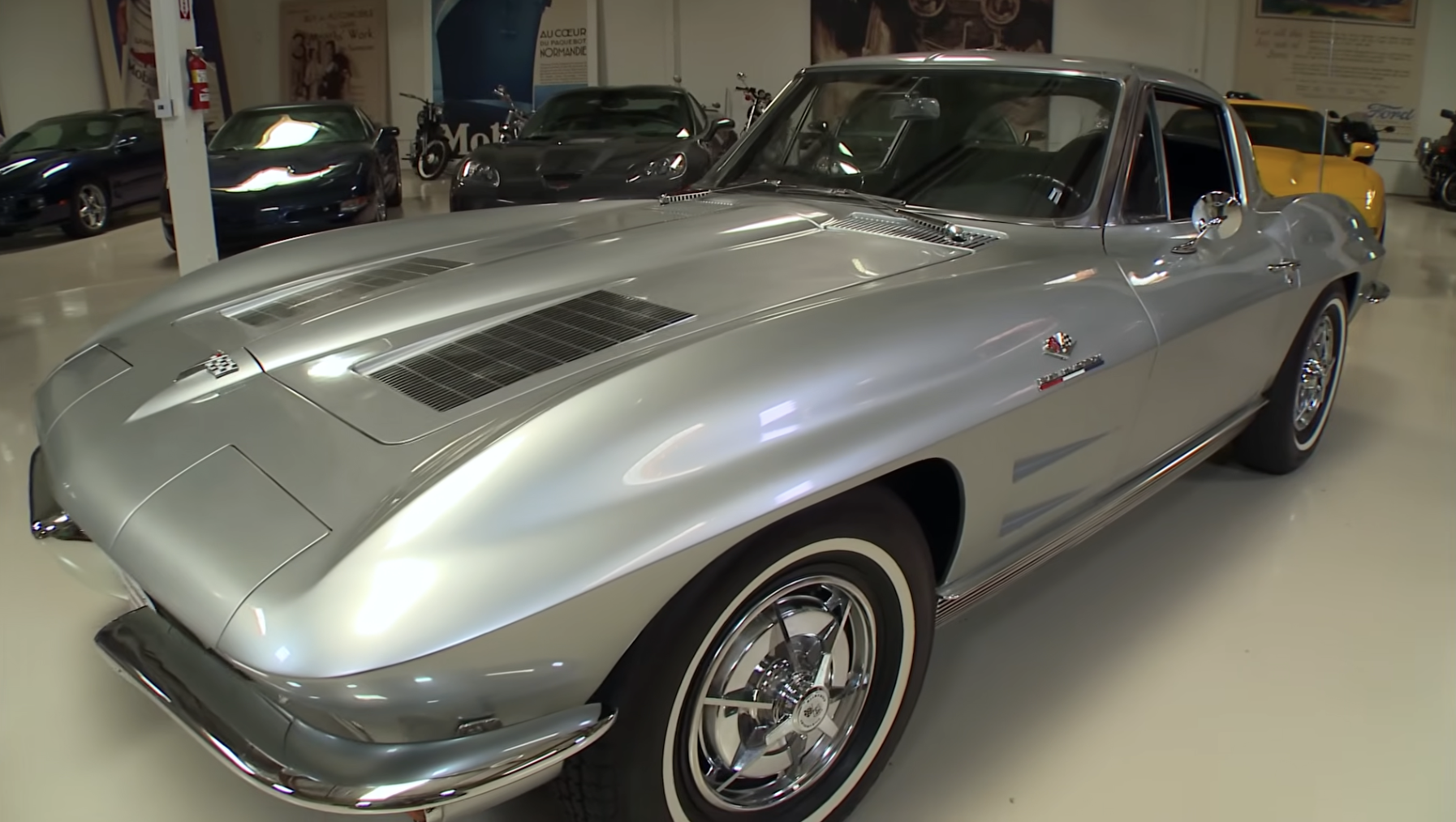A classic silver Chevrolet Corvette Sting Ray sports car is parked indoors under bright lights, with other cars visible in the background.