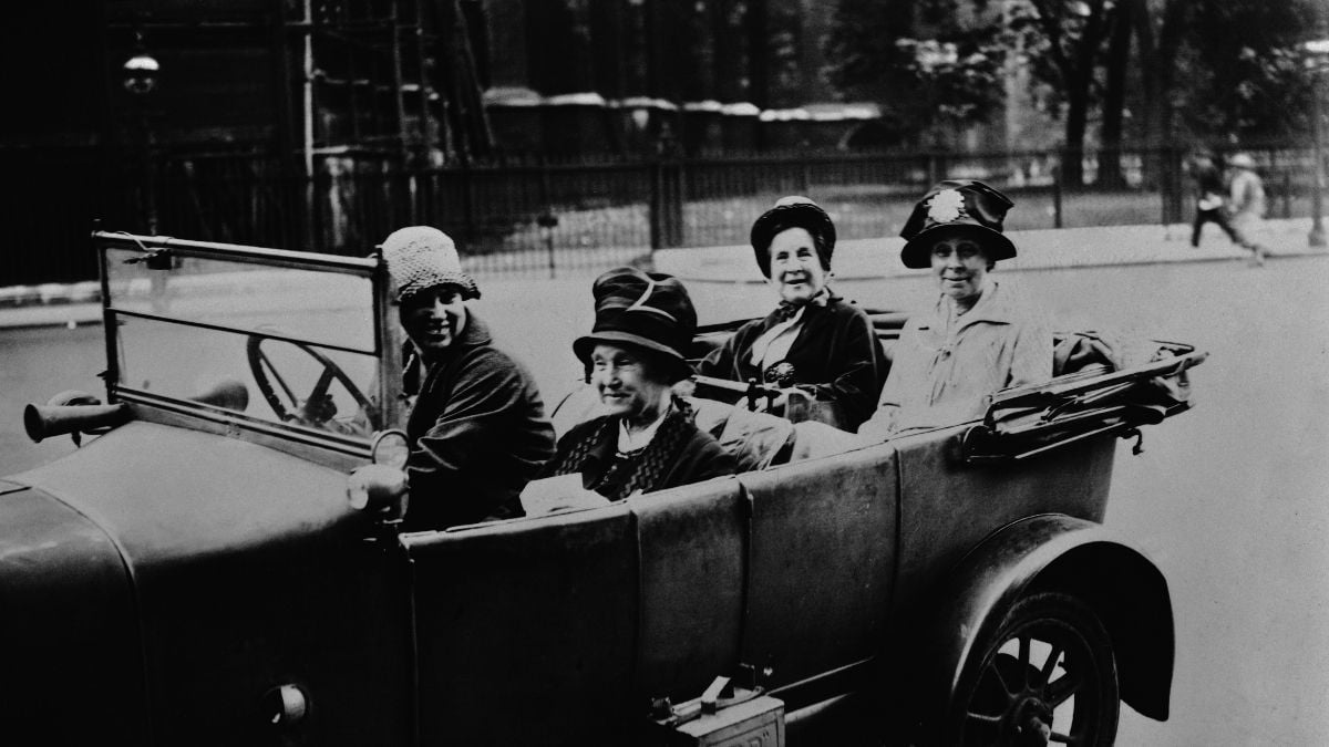 Four women in vintage clothing sit in an open-top car on a city street, smiling for the camera. Trees and a fence are visible in the background. The photo appears to be from the early 20th century.