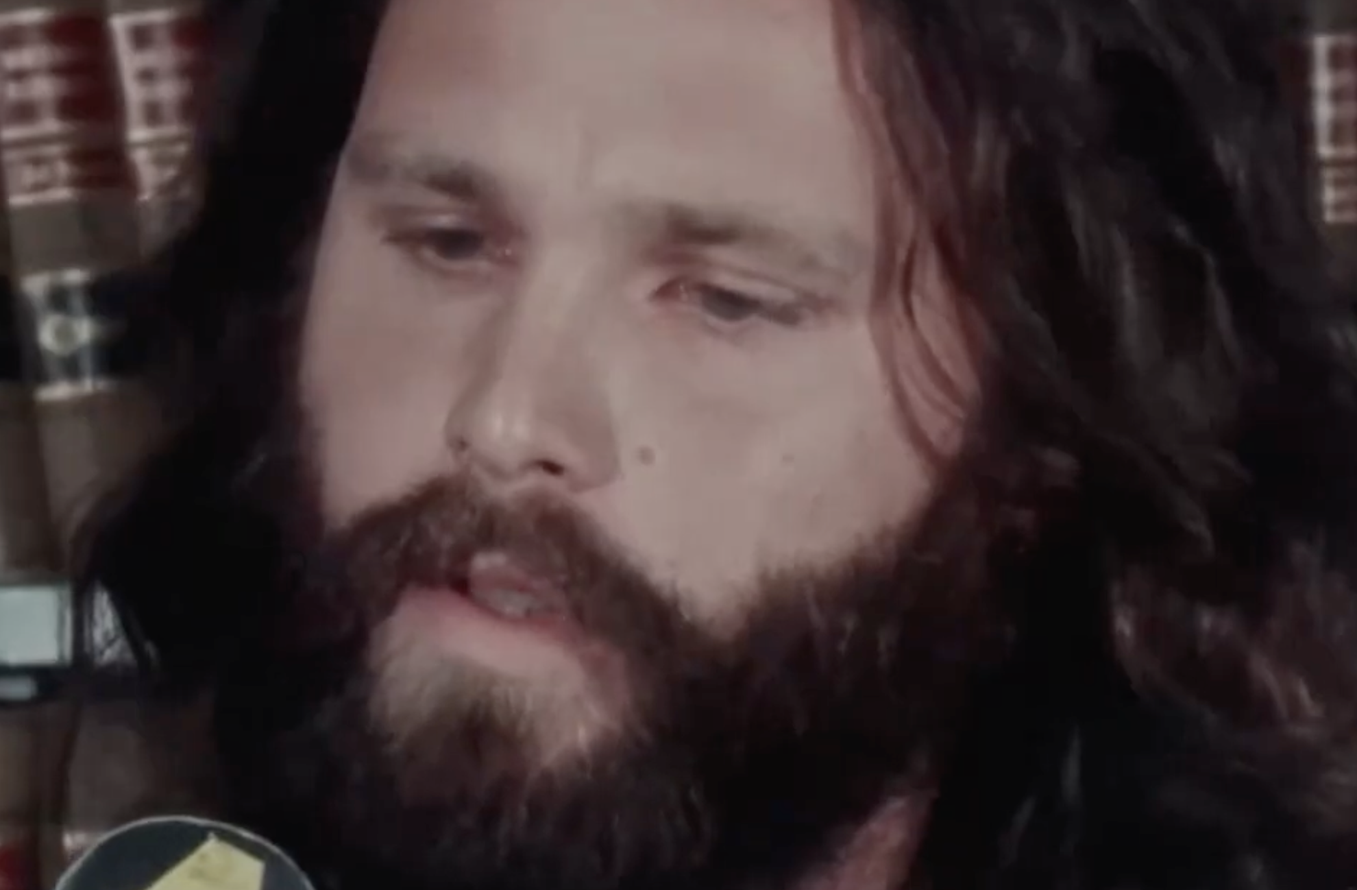 A close-up portrait of Jim Morrison with long dark hair and a beard, speaking into a microphone.