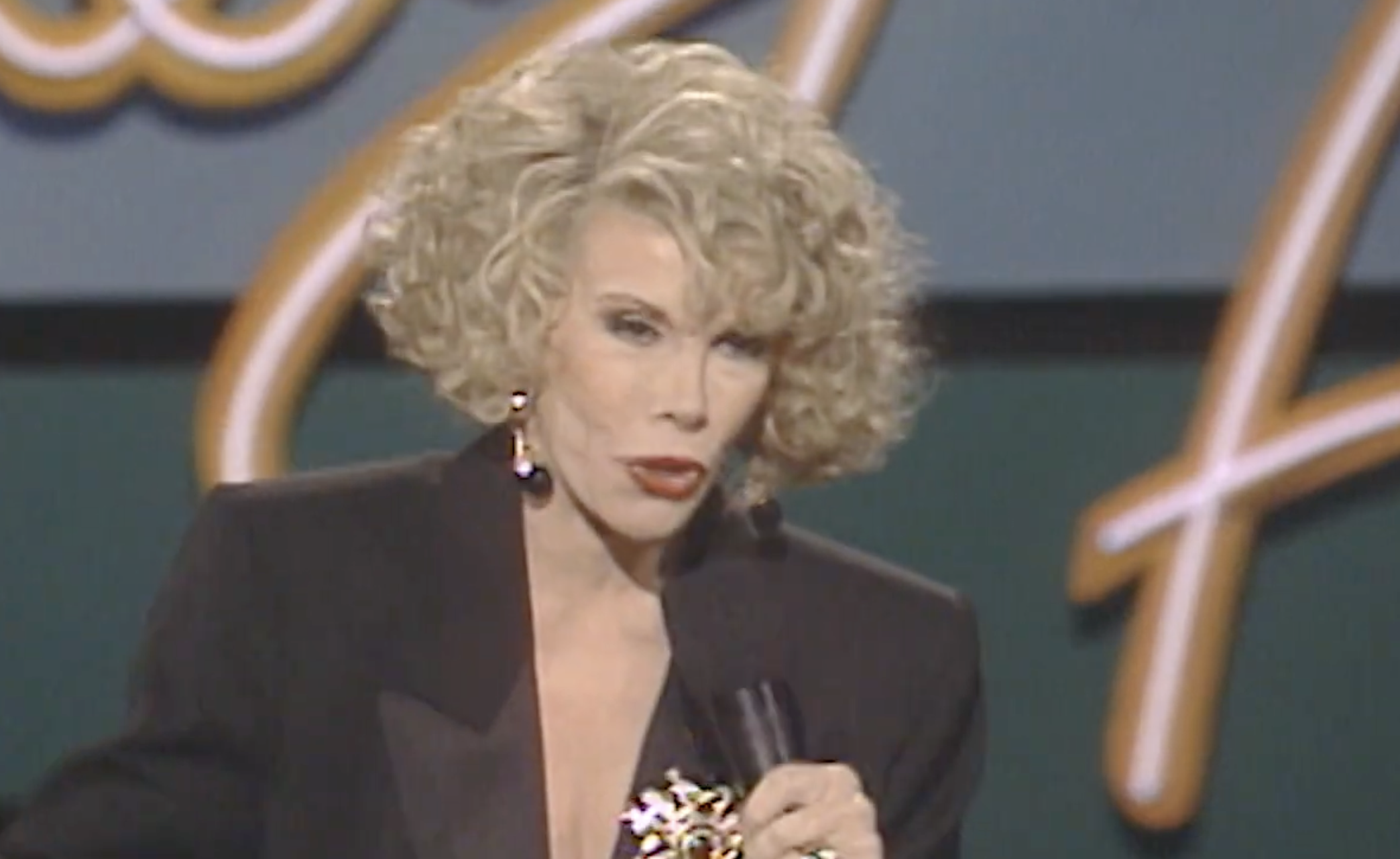 Joan Rivers speaking into a microphone on a stage with a blurred backdrop.