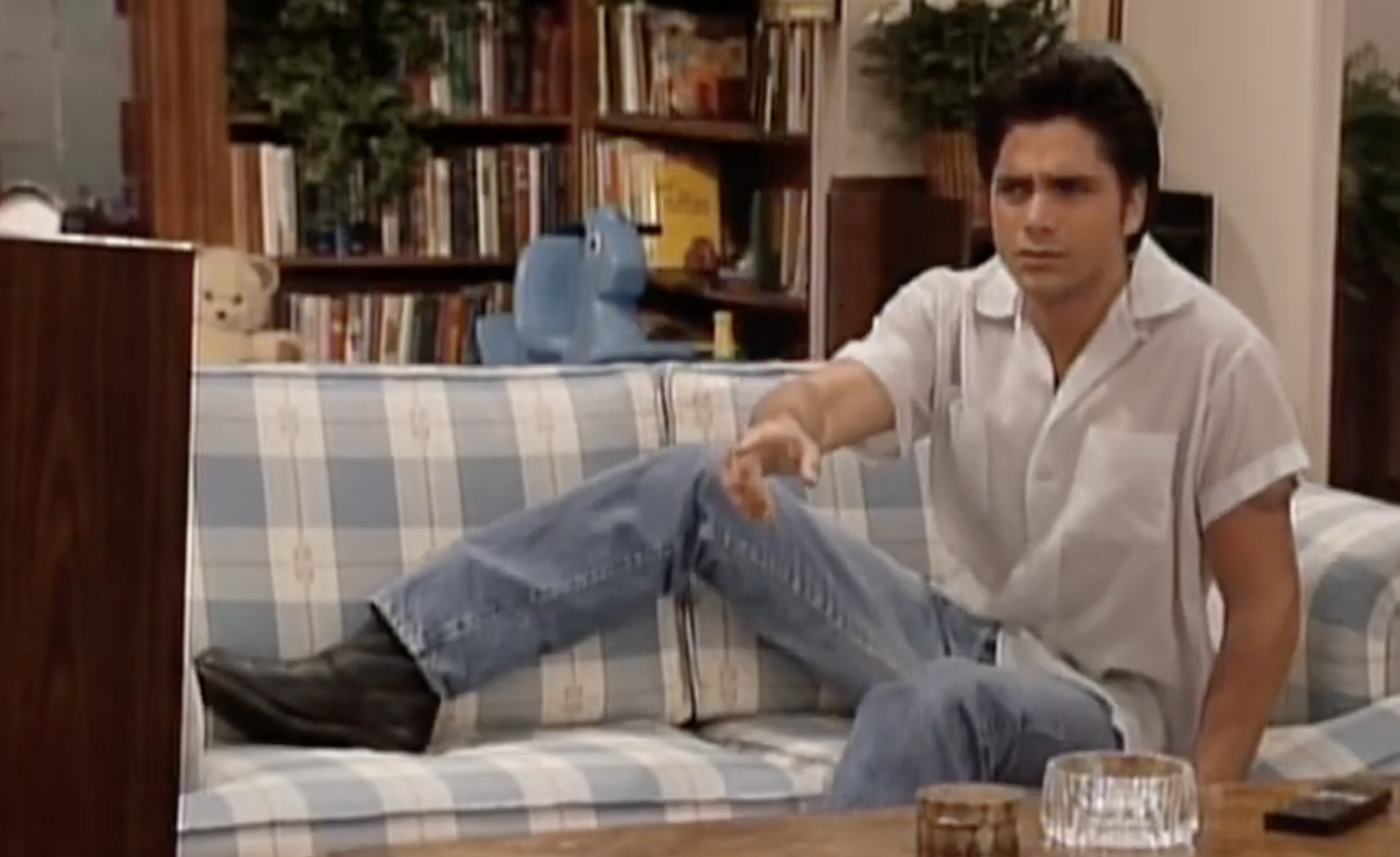 A man sitting on a plaid couch, gesturing toward a television set.