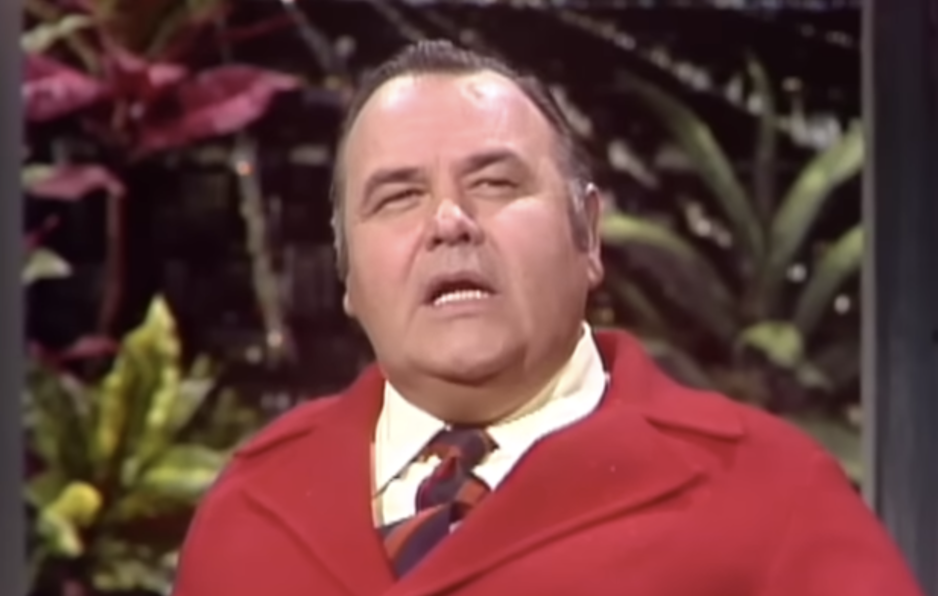 Jonathan Winters wearing a red coat, yellow collared shirt, and a striped tie.