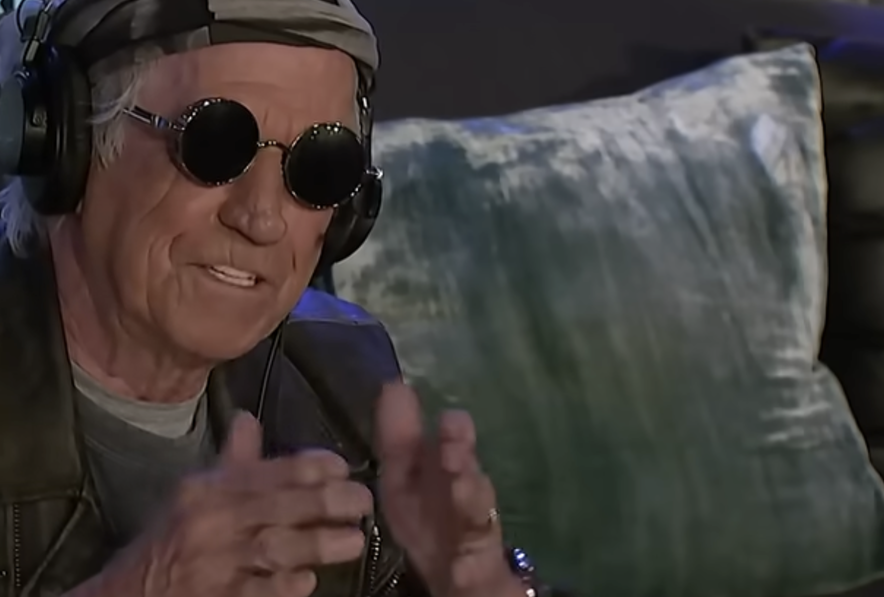 Keith Richards wearing headphones and dark round sunglasses while gesturing with his hands.