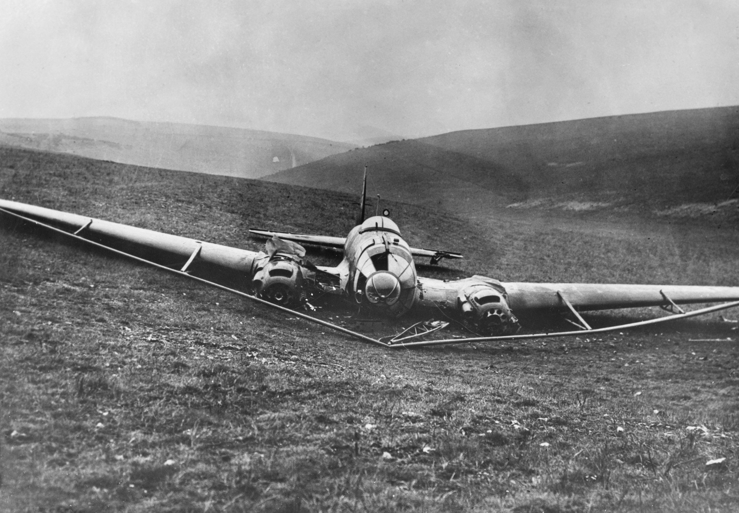 A vintage military airplane with three engines sits crashed on a grassy hillside, its wing bent and damaged. Misty hills are visible in the background.