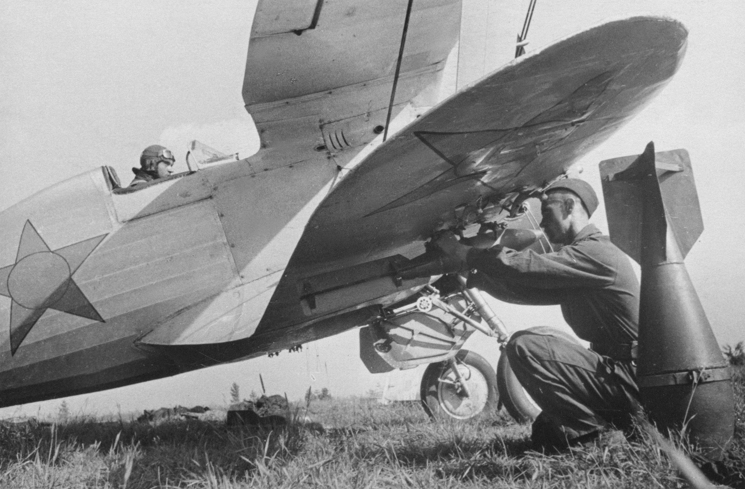 A mechanic works under the tail of a small military aircraft while a pilot sits in the cockpit. The plane is on grass, and a bomb is positioned near the mechanic.