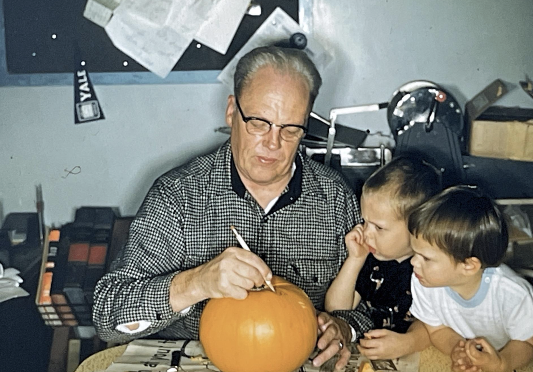 A man wearing glasses carves a pumpkin while two young children watch closely.
