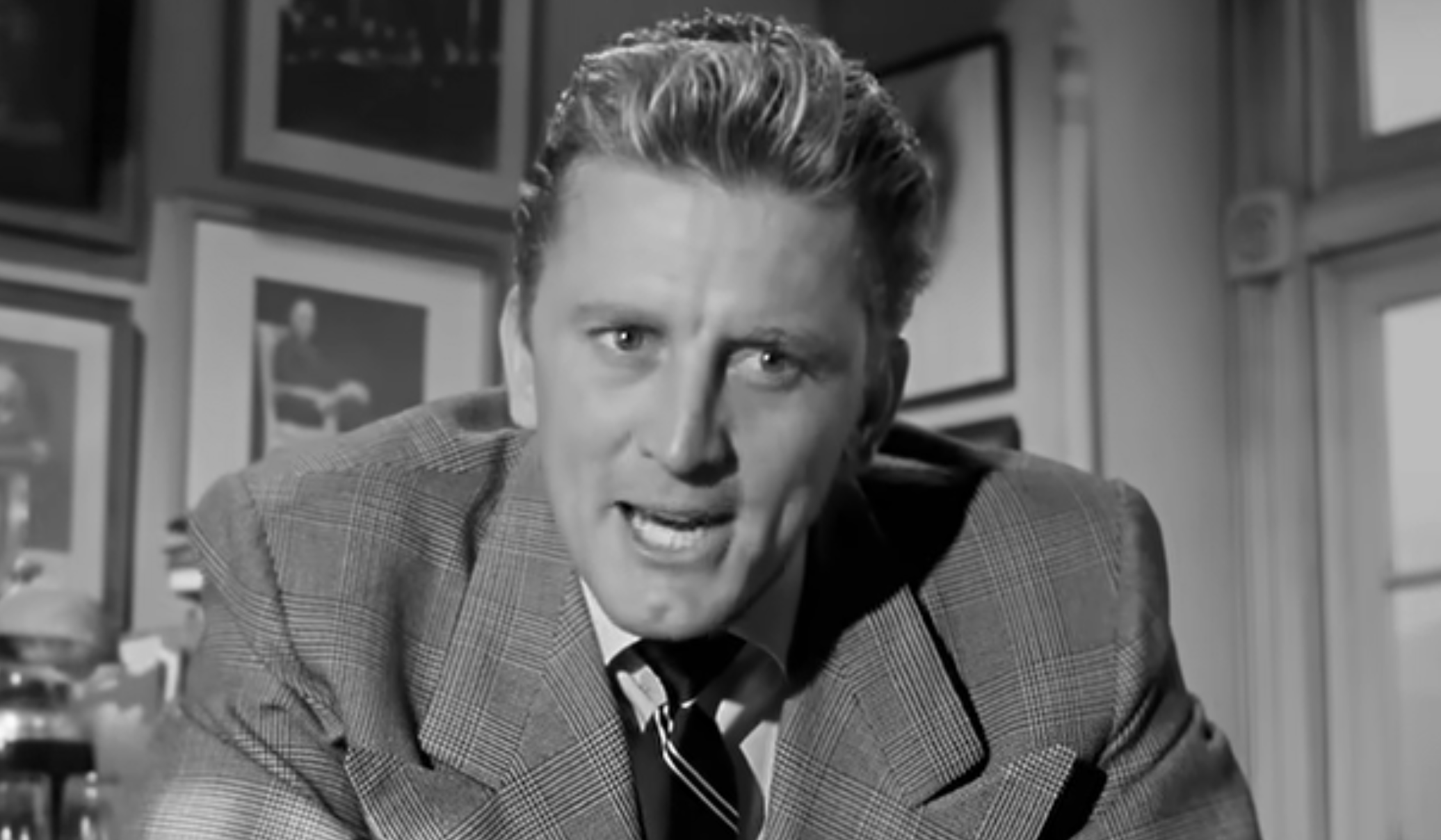 Kirk Douglas in a suit and tie, speaking directly to the camera.