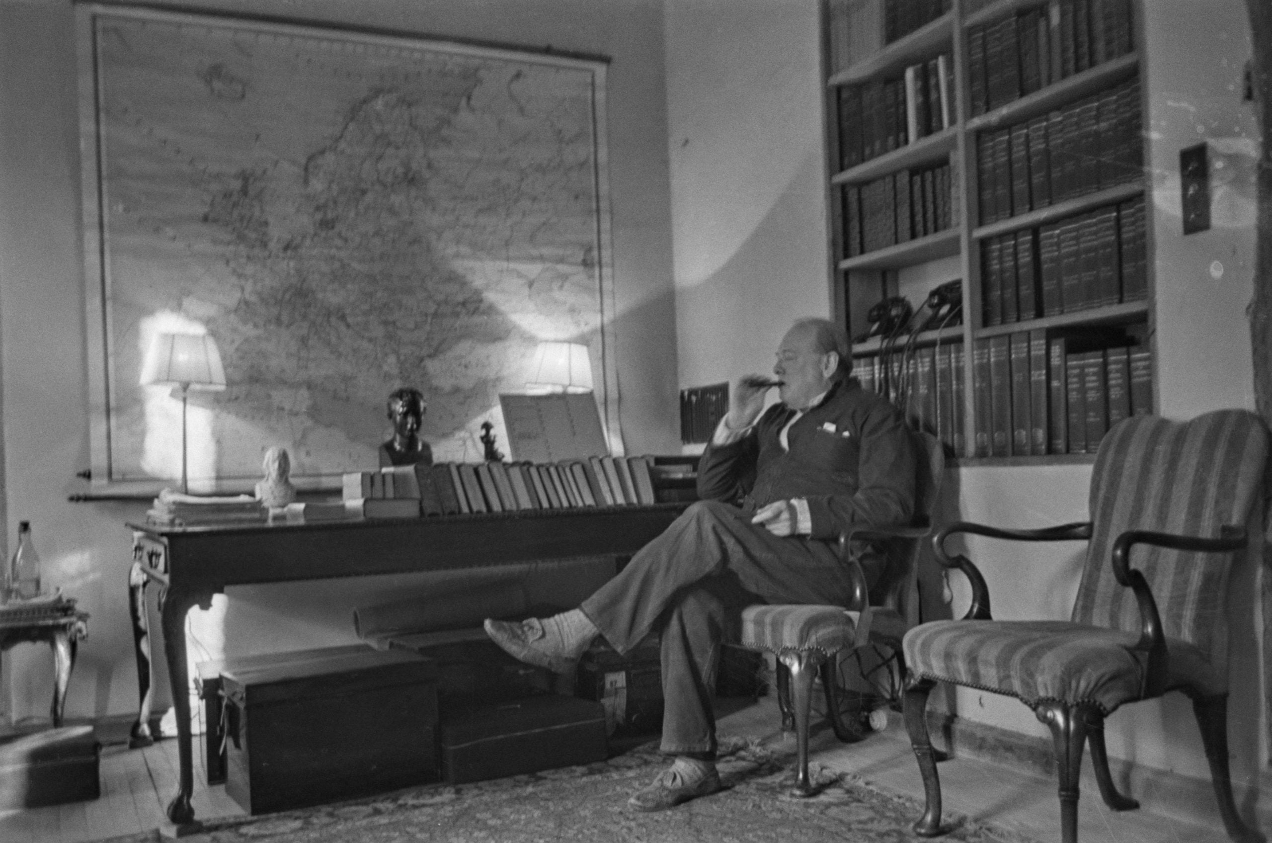 A man sits in an armchair, smoking a cigar in a study with bookshelves, a desk with books, and a large map of Europe on the wall. Two lamps illuminate the room.