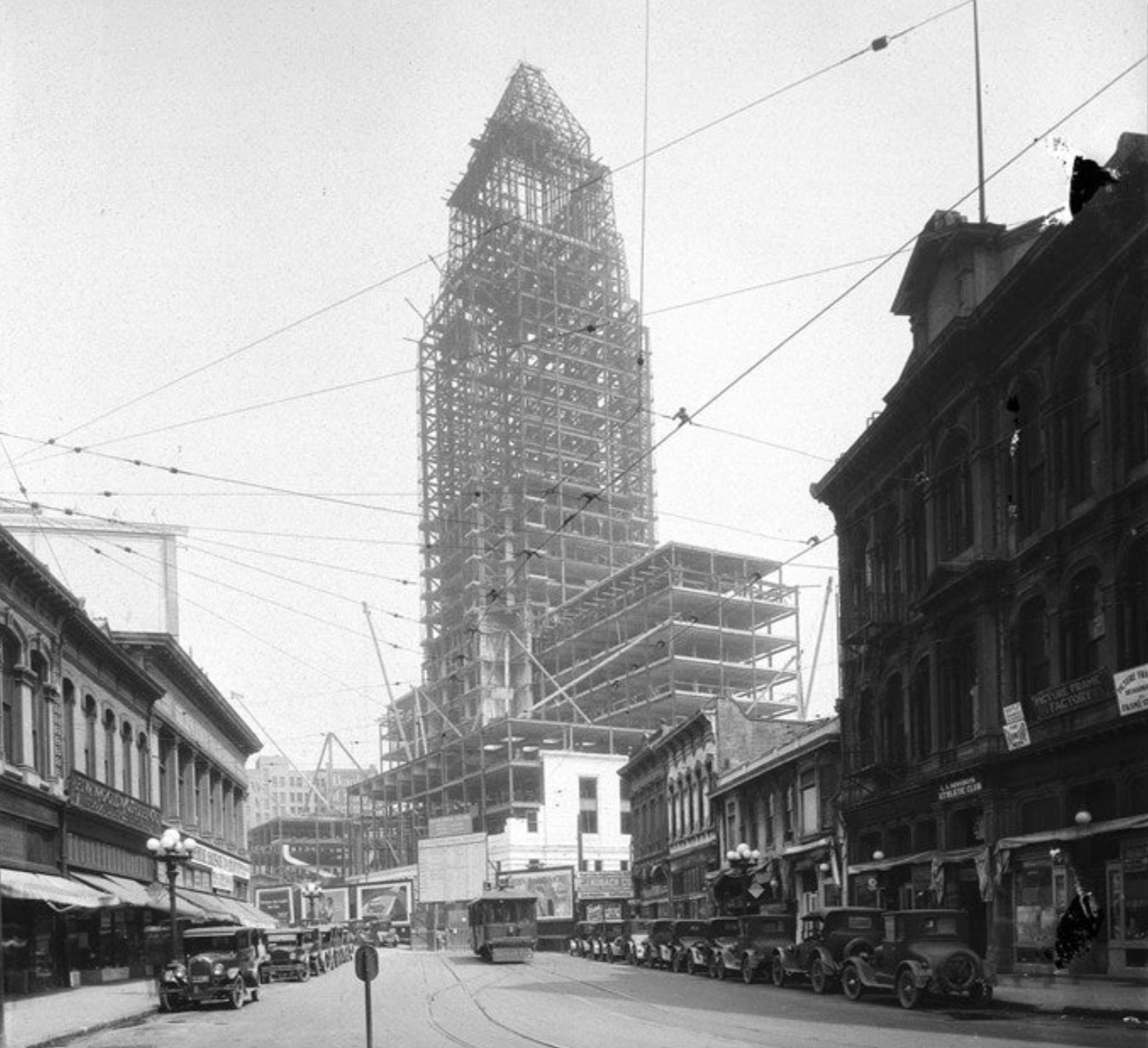 A skyscraper under construction rises above a city street with vintage cars, a streetcar, and surrounding buildings.
