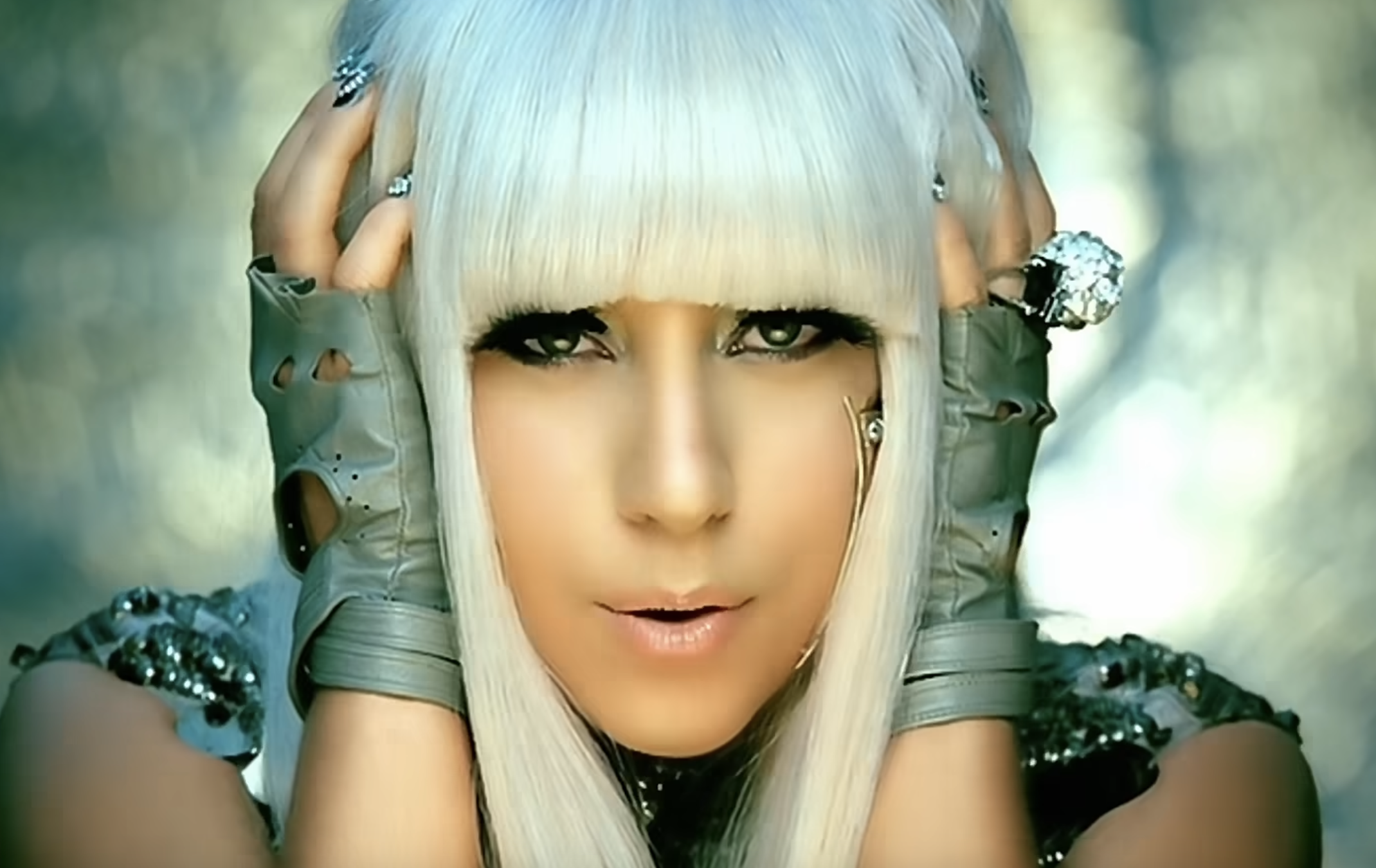 Lady Gaga with platinum blonde hair and bangs, wearing grey fingerless gloves, poses with her hands touching her head.