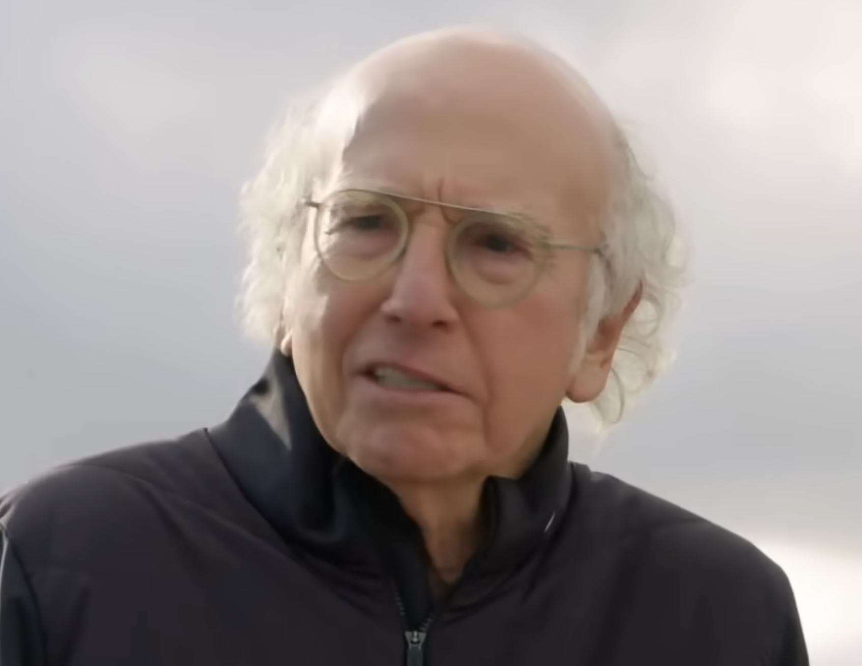 Larry David with a skeptical expression, wearing glasses and a dark zip-up jacket.