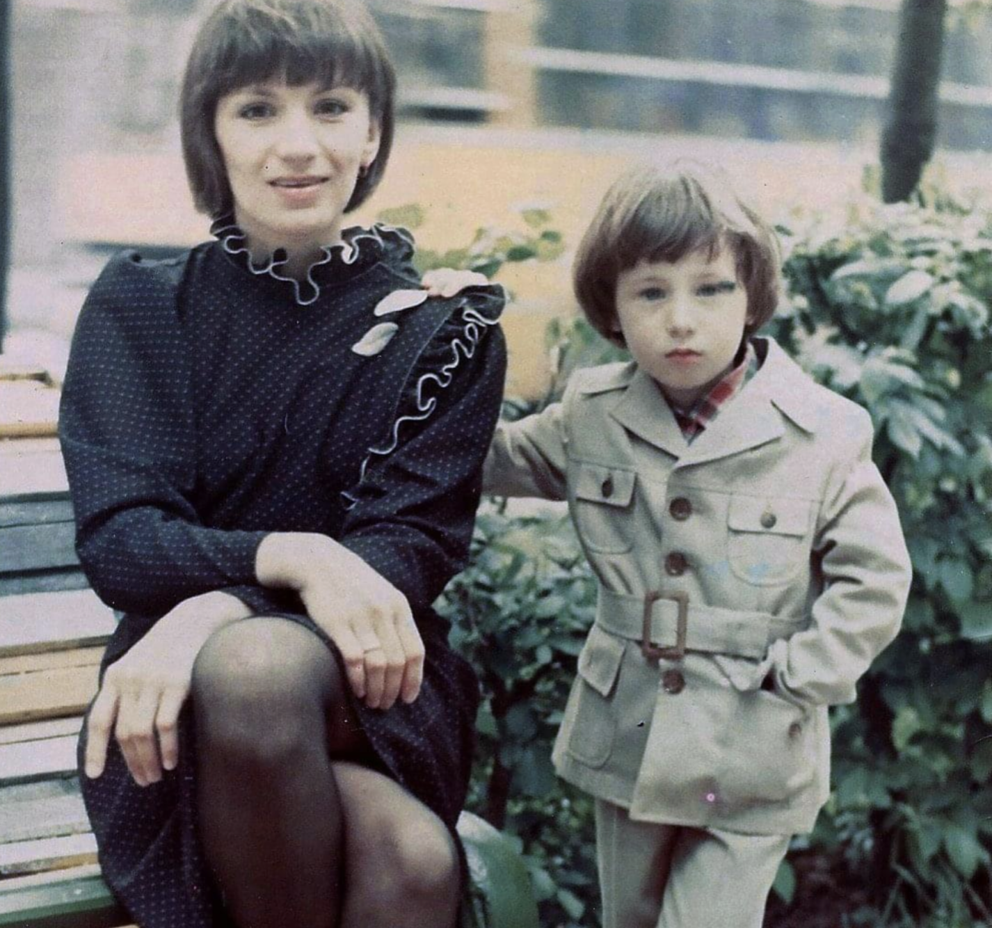 A woman in a dark dress and a young child in a beige suit sitting together on a bench.