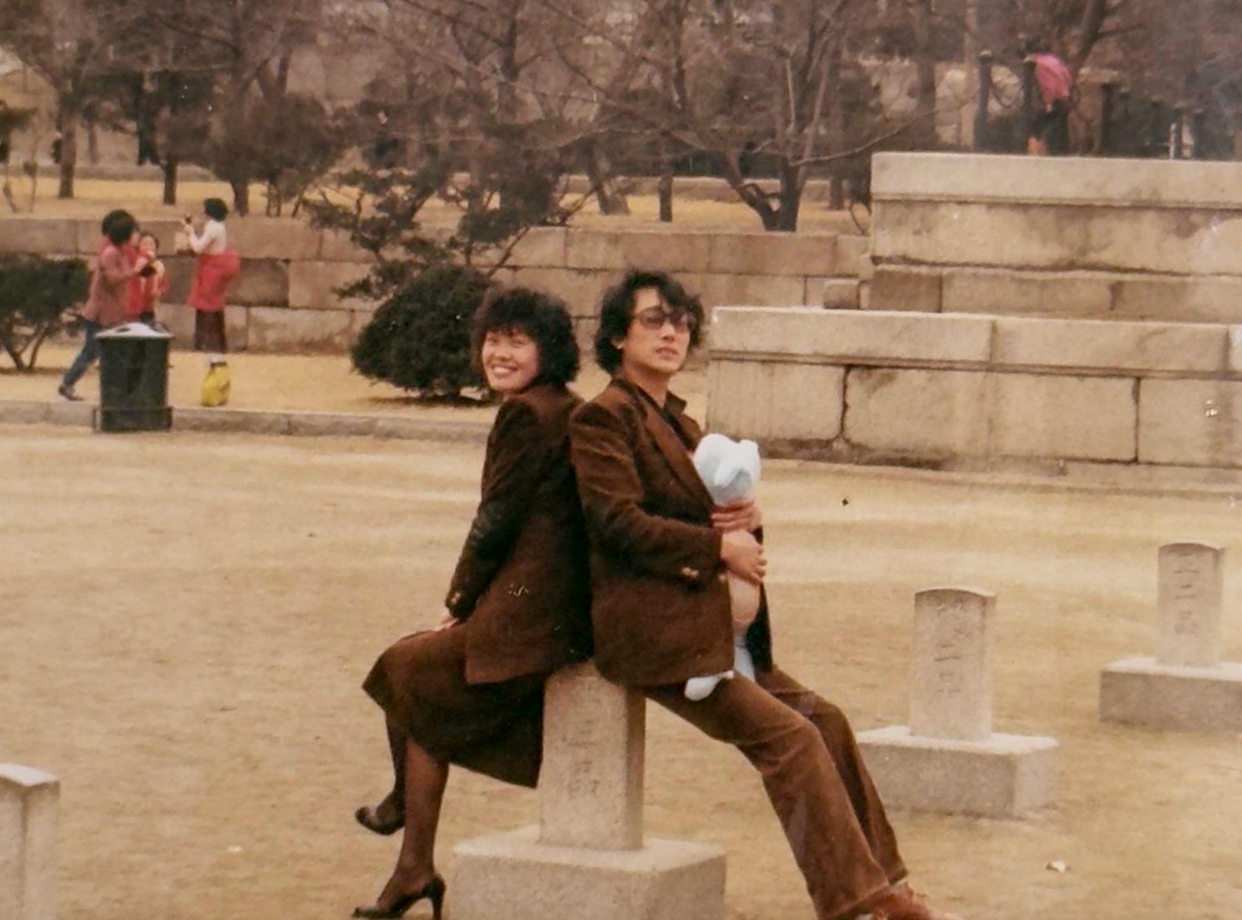A man and woman sitting back-to-back on a stone monument in a park, with other people and stone markers in the background.