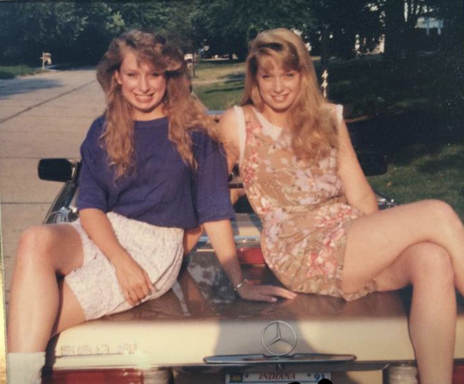 Two young women with long blonde hair sitting on the trunk of a vintage convertible car.