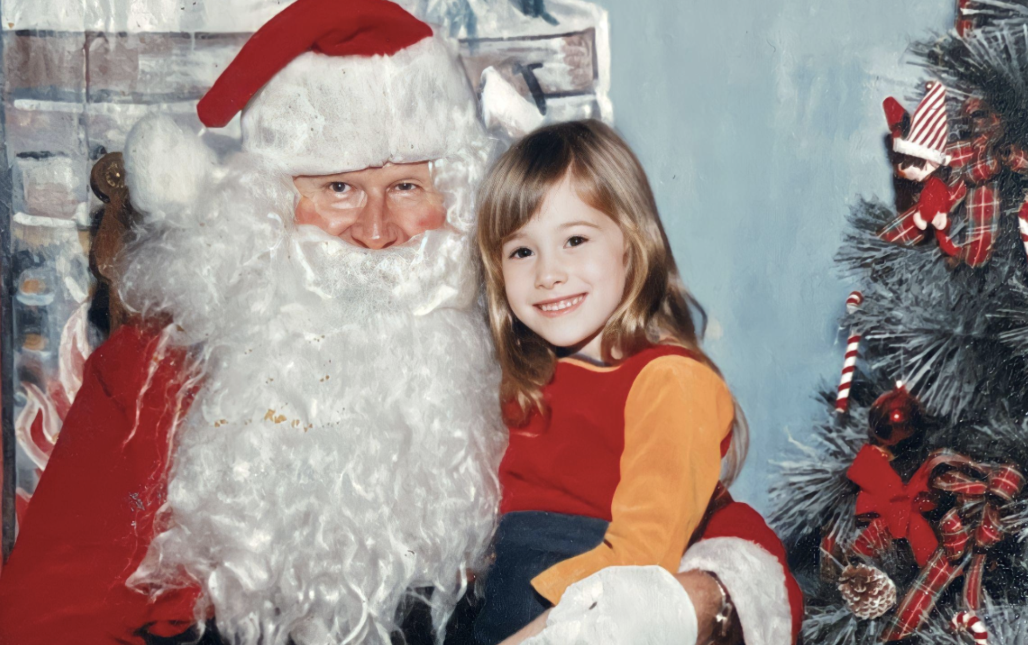 A young girl sits on Santa Claus’s lap next to a decorated Christmas tree.