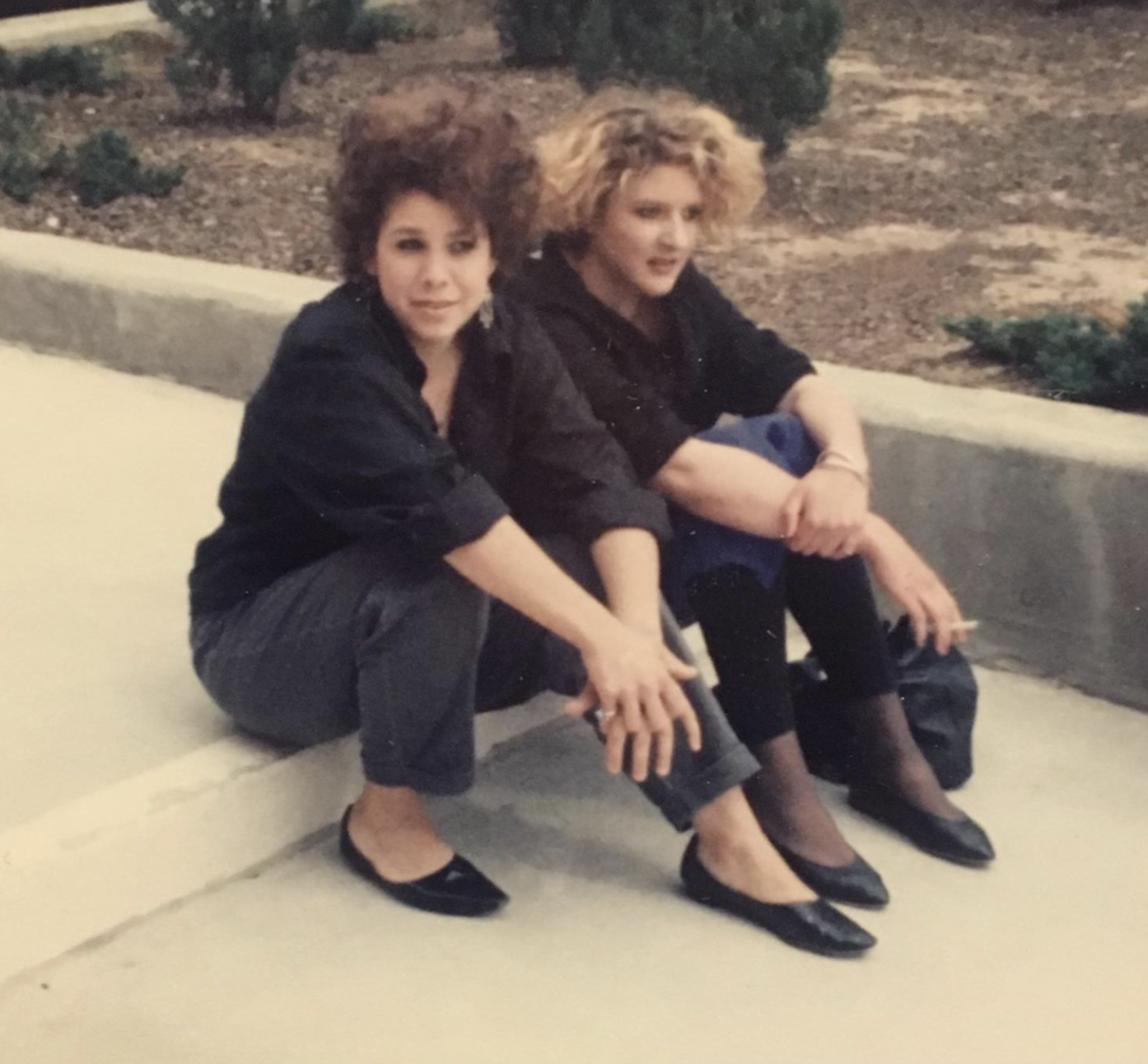 Two women sit on a concrete curb outdoors.
