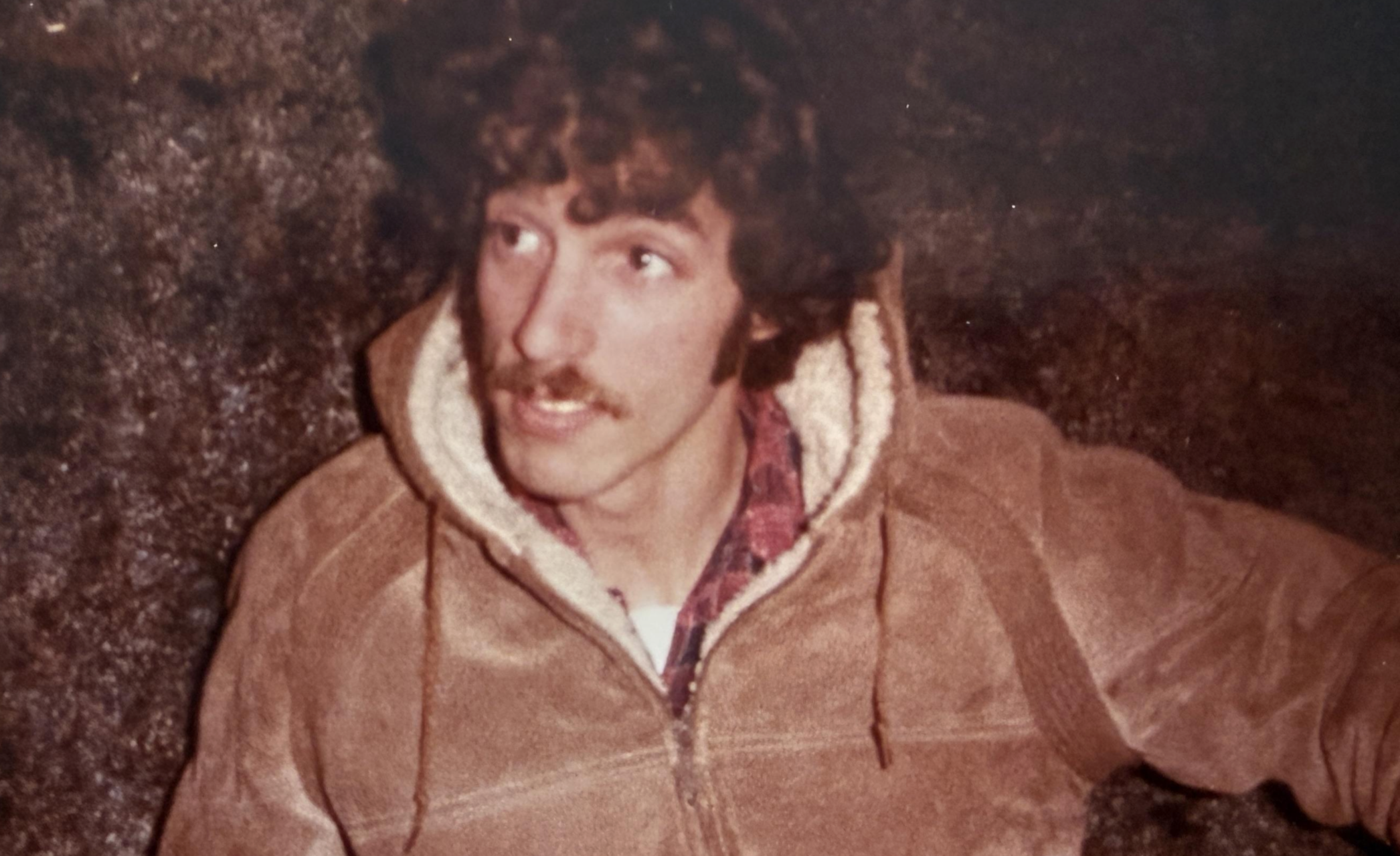A young man with a mustache, wearing a hooded jacket over a plaid shirt, looking off to the side.