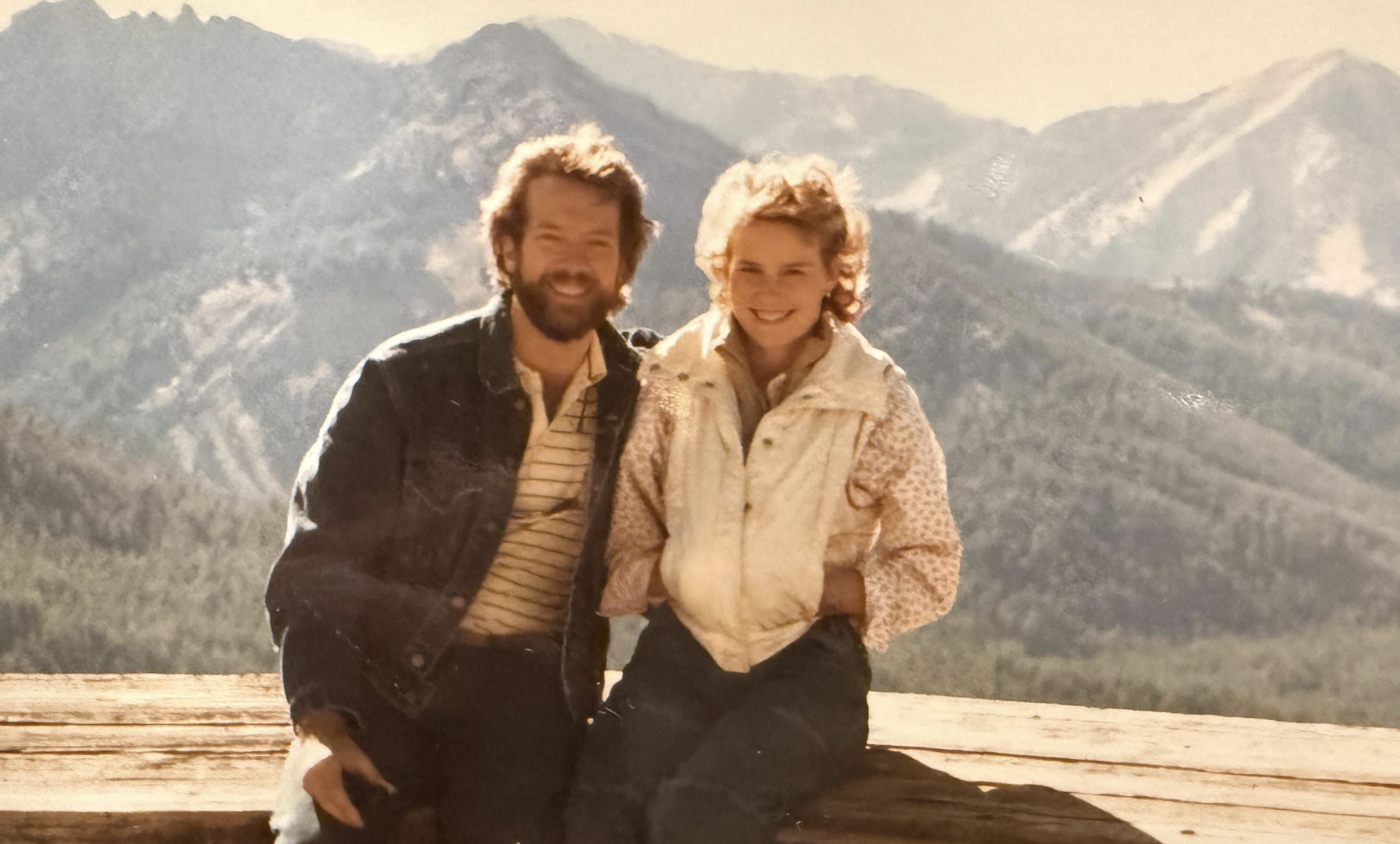 A man and woman smiling while sitting on a wooden platform in front of a mountain range.