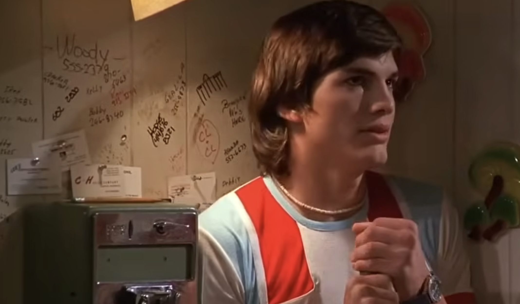 A young man wearing a red, white, and blue ringer t-shirt stands in front of a wall covered in handwritten notes and phone numbers.