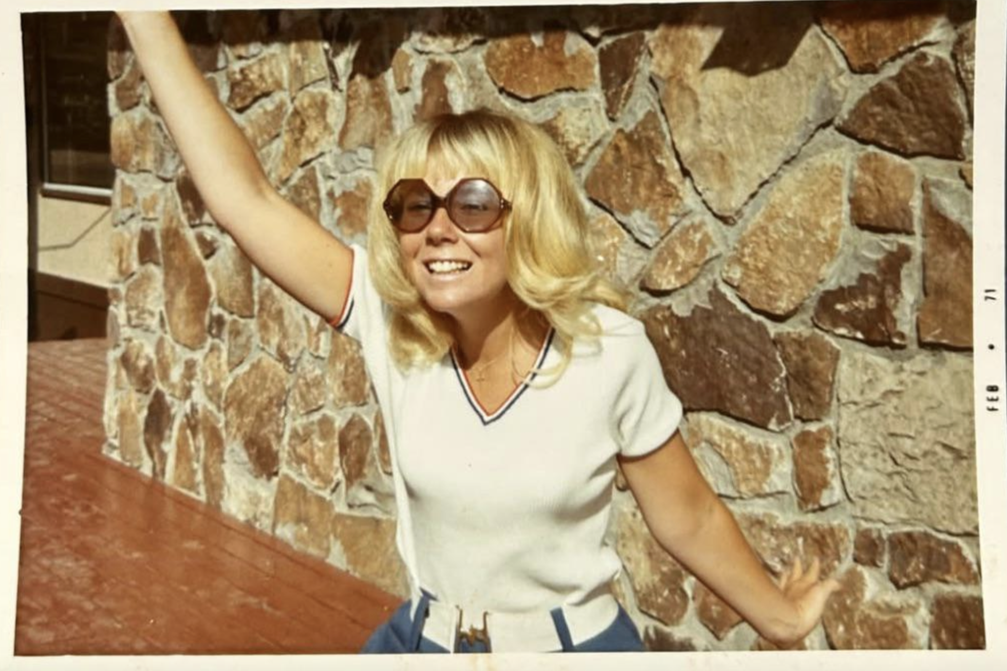 A woman with blonde hair and oversized sunglasses poses with one arm raised in front of a stone wall.