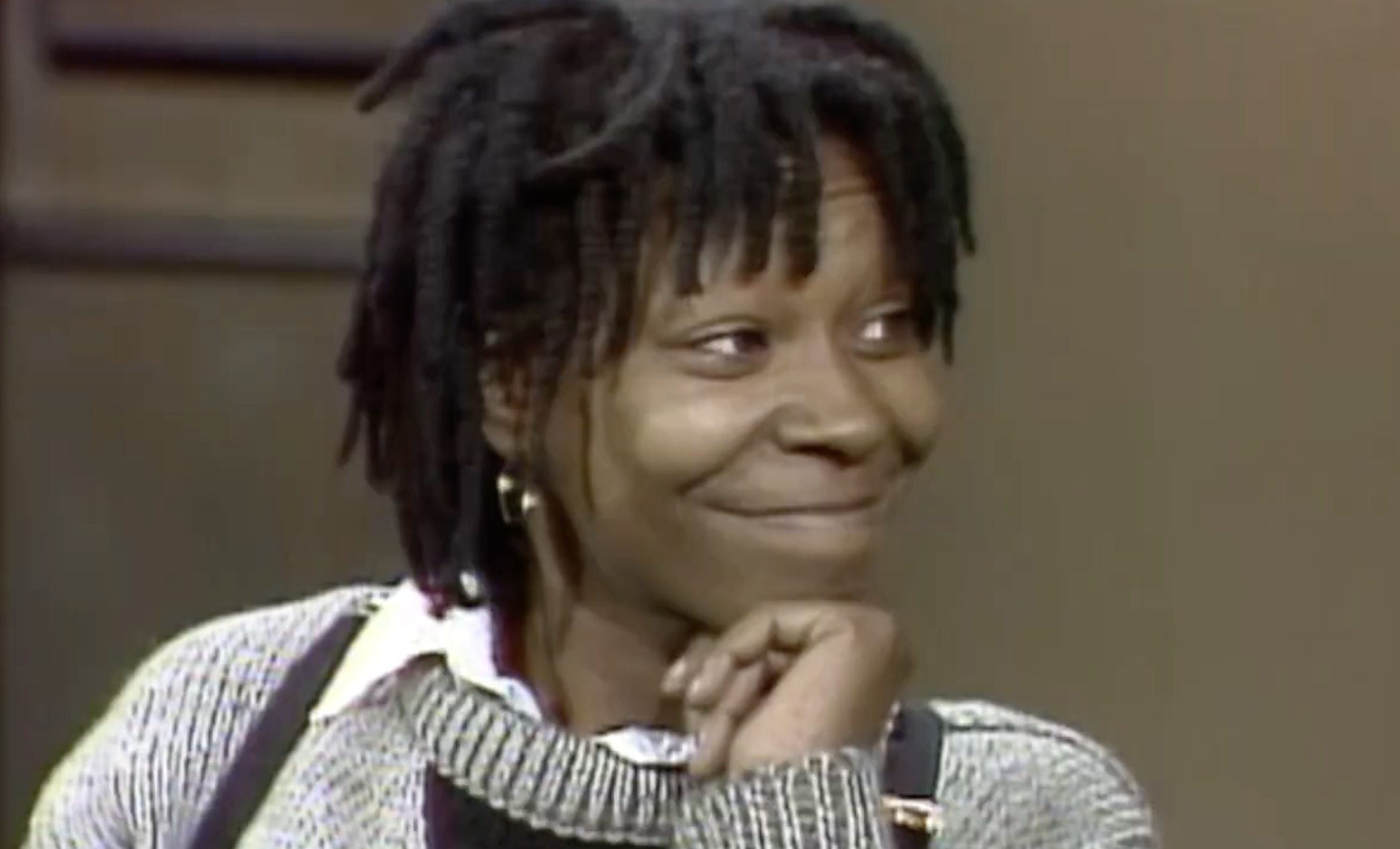 Whoopi Goldberg with dreadlocks, smiling with her hand resting on her chin.
