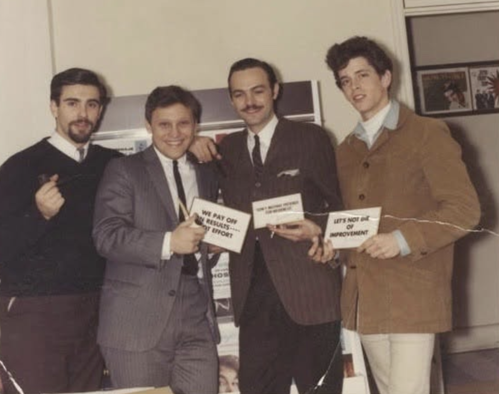 Four men standing side-by-side, three holding signs that read “We pay off in results, not effort,” “I don’t believe in luck, I believe in...” and “Let’s not die of improvement.”